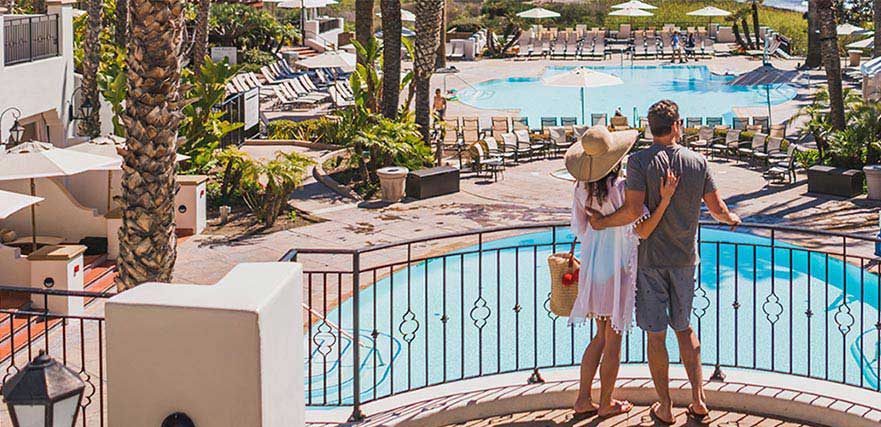 Couple at hotel resort spa, looking out over balcony at two pools and the ocean.