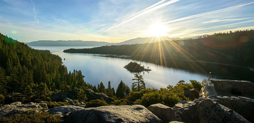 View of valley, lake with small island and the sun shining in the distance past mountainous views.